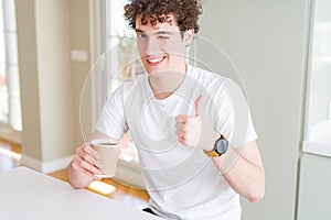 Young man drinking take away cup of coffee at home happy with big smile doing ok sign, thumb up with fingers, excellent sign