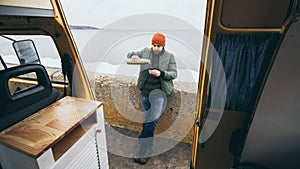 Young man drinking tea from thermos in front of camper van with a lake on background