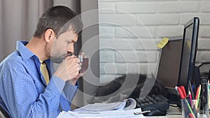 A young man drinks tea from a mug, works at home, a cat lies and yawns in the background