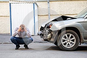 Young man driver in car accident holding his head near broken car on the road after car accident. Caucasian man facepalm