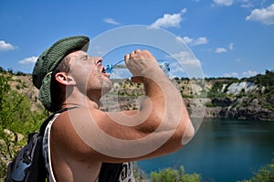 A young man with drops of sweat on his face, in a hat and with a backpack drinks cool clean water from a plastic bottle on a hot s