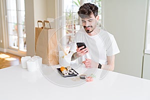 Young man eating asian sushi from home delivery and ordering food using smartphone app with a happy face standing and smiling with