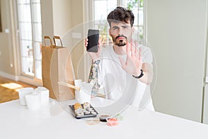 Young man eating asian sushi from home delivery and showing smartphone screen with open hand doing stop sign with serious and