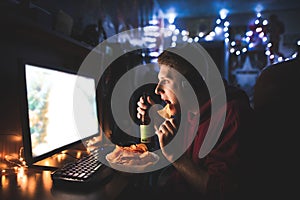 Young man eats chips, drinks beer and looks at the computer screen, sits at the table at home in a cozy room