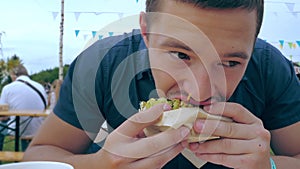 A young man eats a hamburger or cheeseburger with a green bun in an open-air cafe at a festival or village fair.