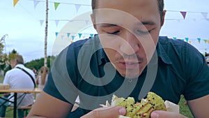 A young man eats a hamburger or cheeseburger with a green bun in an open-air cafe at a festival or village fair.