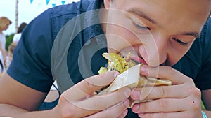 A young man eats a hamburger or cheeseburger with a green bun in an open-air cafe at a festival or village fair.