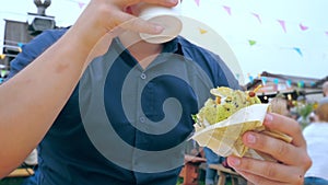 A young man eats a hamburger or cheeseburger with a green bun in an open-air cafe at a festival or village fair.