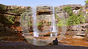 Young man enjoying the pristine natural waterfall falling from mountain top at day from low angle