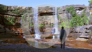 Young man enjoying the pristine natural waterfall falling from mountain top at day from low angle