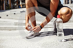 Young man exercising outside. Cut low view of man tie laces of shoes or sneakers. Almost empty water bottle besides on