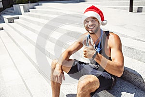 Young man exercising outside. Guy wearing christmas santa hat and show big thumb up. Like symbol. Strong powerful
