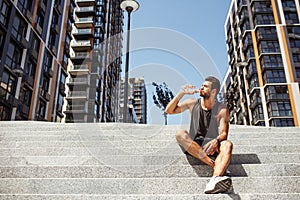 Young man exercising outside. Picture of athletist drinking water ad sitting on steps outside. Urban view. Body