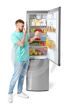 Young man with expired sausage near open refrigerator on white background