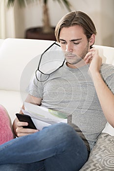 Young man with a face mask hanging from his right ear reading some papers and holding his phone