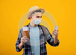 Young man in face mask holding passport and tickets, using smartphone to book vacation online, orange background