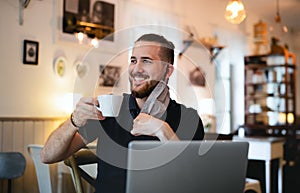 Young man with face mask and laptop indoors in cafe, drinking coffee.