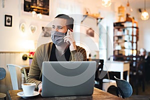 Young man with face mask and laptop indoors in cafe, using smartphone.