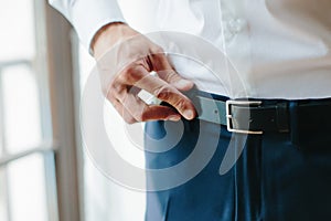 Young man fastens black belt on his trousers, male hands close up. Morning of groom, preparations for wedding. Selective focus.