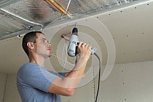 Young man fixing drywall suspended ceiling to metal frame using electrical screwdriver.