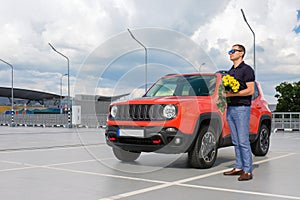 A young man with flowers in his hands standing near his car.