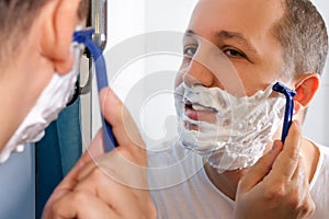 Young man with foam on his face shaves in front of a mirror.