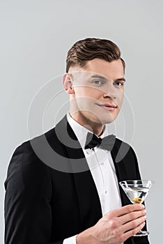 Young man in formal wear with bow tie holding glass of cocktail isolated on grey