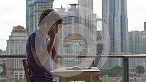 Young man freelancer workes on his laptop at a balcony with a background of a city center full of skyscrapers. Remote