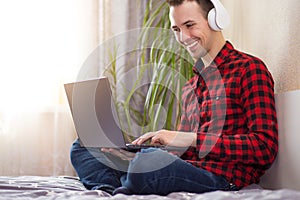 Young man freelancer working with laptop at home in bed near window and listen to music in headphones. Handsome man reads on