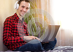 Young man freelancer working with laptop at home in bed near window and listen to music in headphones. Handsome man reads on