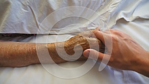 Young man funny takes and comforts hand of his elderly grandmother in hospital. Grandson gently touches wrinkled arm of