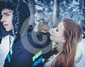 Young man in a fur hat with a earflap with a kitten in the hood of his jacket and a girl in a beige fur coat against the