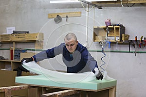 Young man in a furniture factory applies a foam on a wooden piece of the sofa