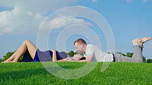 The young man gently kisses his pregnant wife. Together lie on a green lawn in a park against a blue sky