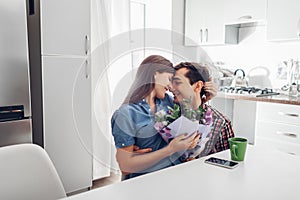Young man gifting bouquet of flowers to his girlfriend in kitchen. Happy couple hugging. Romantic surprise