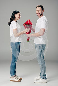 Young man gifting bouquet of red roses to pretty girlfriend on grey background