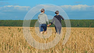 A young man and a girl are holding hands walking along a cereal field.