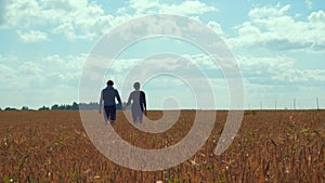 A young man and a girl are holding hands walking along a cereal field.