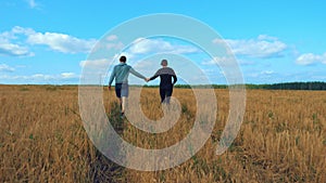 A young man and a girl are holding hands walking along a cereal field.