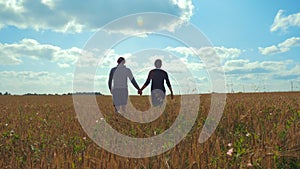 A young man and a girl are holding hands walking along a cereal field.