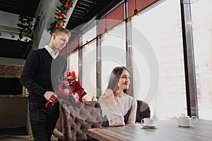 A young man gives a bouquet of red flowers to his girlfriend, wife, in a cafe by the window.