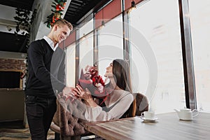 A young man gives a bouquet of red flowers to his girlfriend, wife, in a cafe by the window.