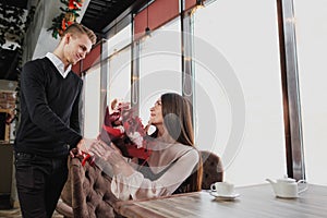A young man gives a bouquet of red flowers to his girlfriend, wife, in a cafe by the window.