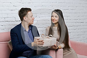 A young man gives a gift to a girl. He hands her a box of wrapping paper tied with a ribbon. Sitting on a sofa against a white