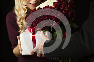 A young man gives a gift a white box with a red bow and flowers to a girl on an isolated black background. Valentine day concept