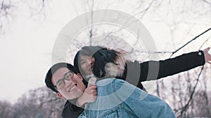 Young man giving piggyback ride to his girlfriend in the park on snowy winter day. Newlywed asian couple having fun