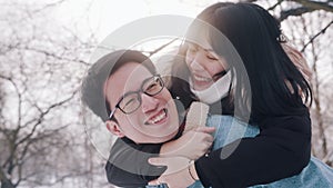 Young man giving piggyback ride to his girlfriend in the park on snowy winter day. Newlywed asian couple having fun