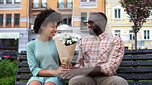 Young man giving white flower bouquet to woman sitting bench, first awkward date