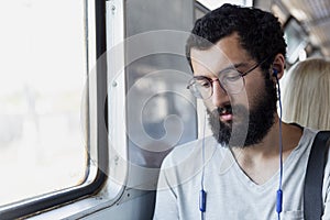 A young man with glasses, headphones and a beard sits in a train car and listens to music. Tourism and travel. Close-up