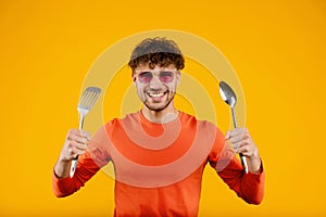 Young man in glasses holds kitchen spatula in one hand and spoon in another , isolated yellow background